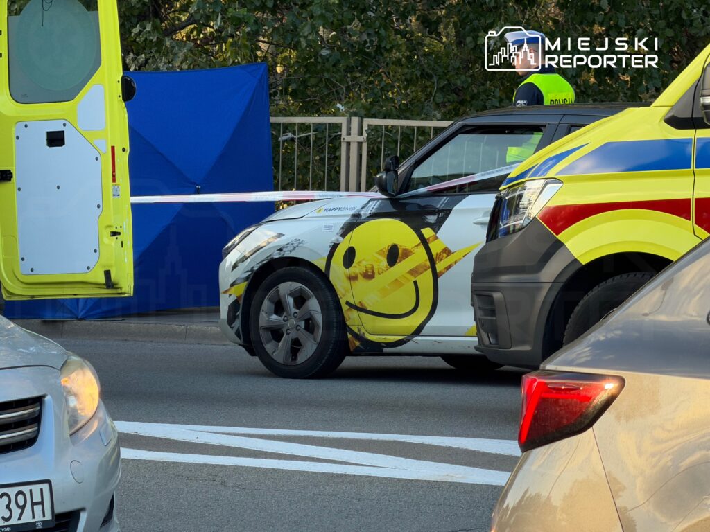 Karetka i samochód z żółtym symbolem uśmiechniętej twarzy stoją obok odgrodzonego niebieskim parawanem miejsca zdarzenia.