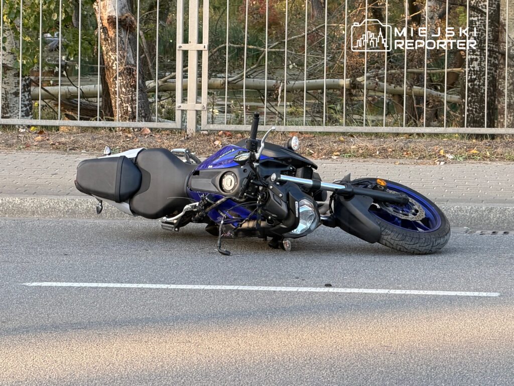 Niebiesko-czarny motocykl leży przewrócony na jezdni przy metalowym ogrodzeniu.