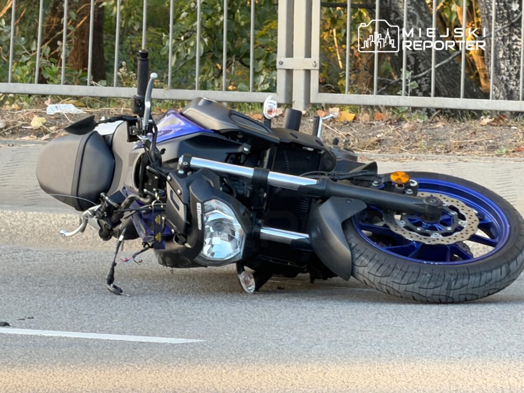 Motocykl leżący na boku na jezdni przy metalowym ogrodzeniu.
