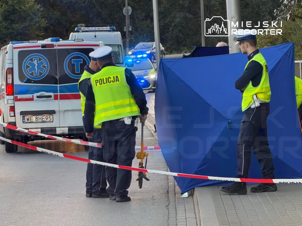 Policjanci zabezpieczają taśmą ulicę przy karetce i niebieskim parawanie rozstawionym na chodniku.