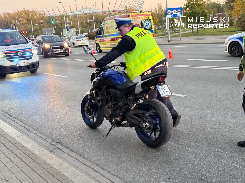 Policjant w odblaskowej kamizelce prowadzi motocykl przez skrzyżowanie, obok stoją karetka i samochody.