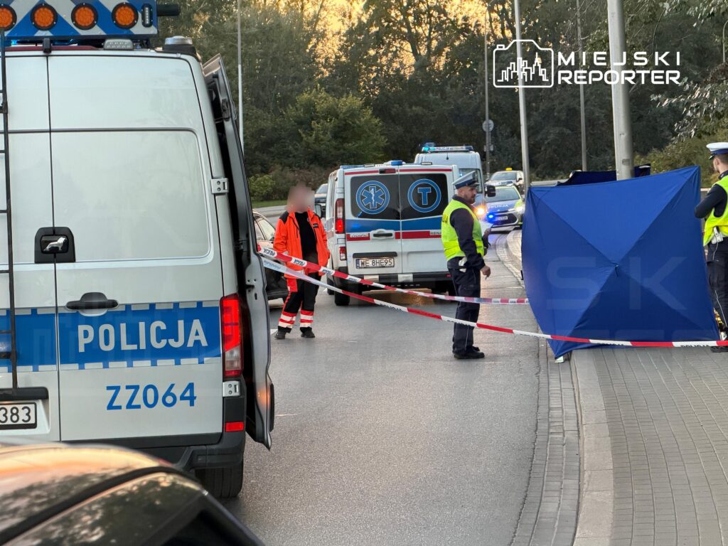 Policjanci i ratownicy zabezpieczają taśmą miejsce zdarzenia na ulicy, obok radiowozu, karetki i niebieskiego parawanu.