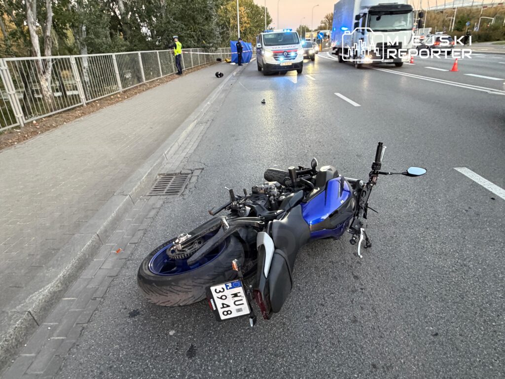 Przewrócony niebiesko-czarny motocykl leży na jezdni, a w tle stoi radiowóz i ciężarówka.
