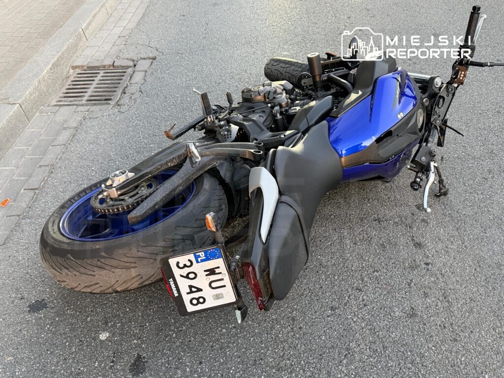 Niebieski motocykl leży przewrócony na jezdni, obok krawężnika i kratki ściekowej.