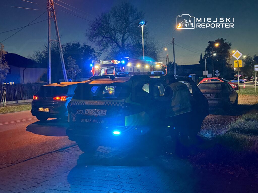 Radiowóz straży miejskiej z włączonymi niebieskimi światłami stoi nocą przy ulicy, obok innych pojazdów służbowych.