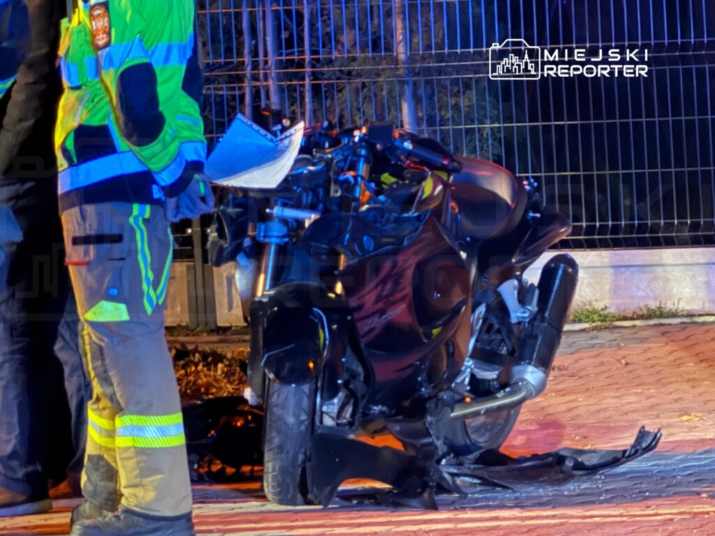 Motocykl leży przewrócony na jezdni, obok stoi strażak w odblaskowym ubraniu ochronnym.