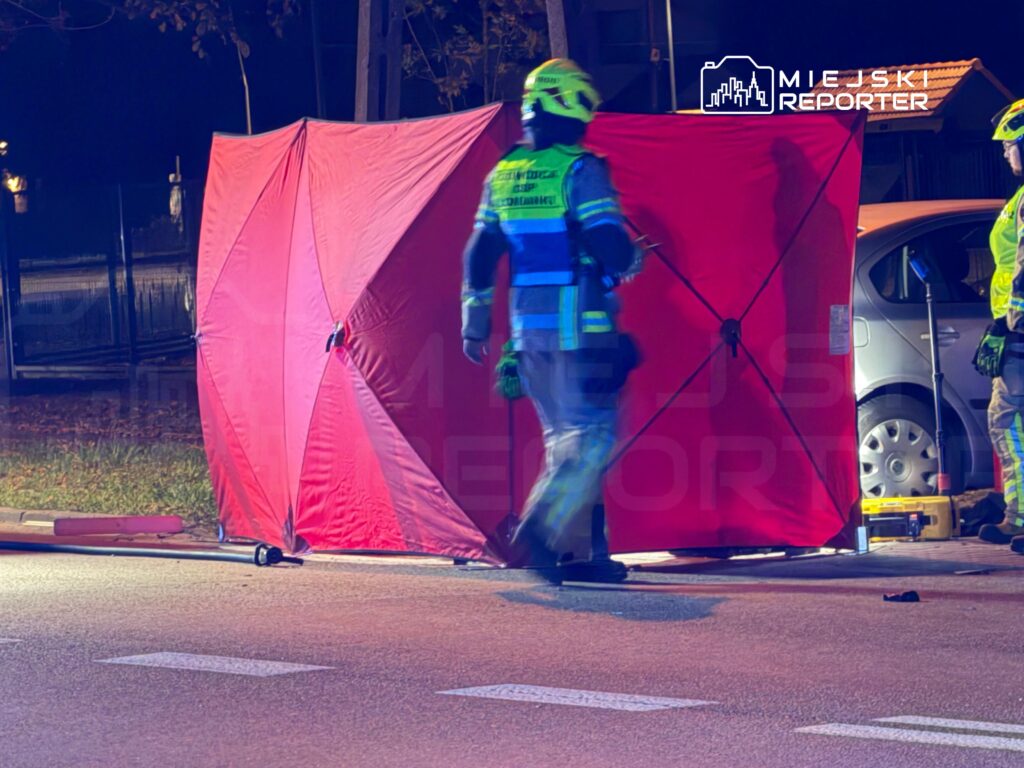Ratownik w odblaskowym stroju przechodzi obok czerwonego parawanu osłaniającego miejsce zdarzenia na nocnej ulicy.