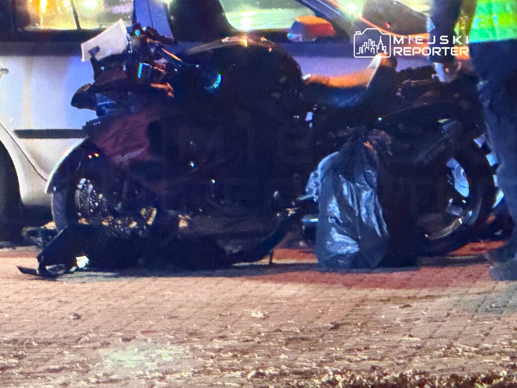 Rozbity motocykl leży na jezdni obok zaparkowanego samochodu, nocą w świetle ulicznych lamp.