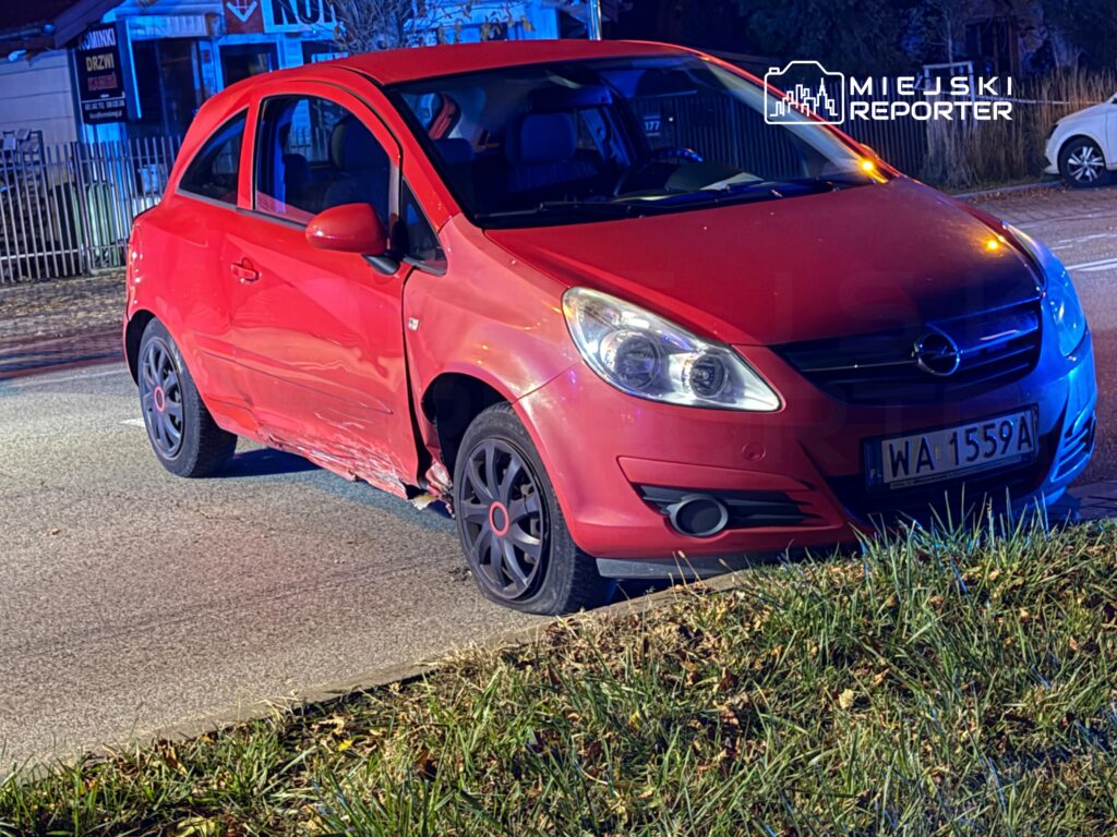 Czerwony Opel z uszkodzonym bokiem stoi przy krawędzi drogi, częściowo na trawniku.
