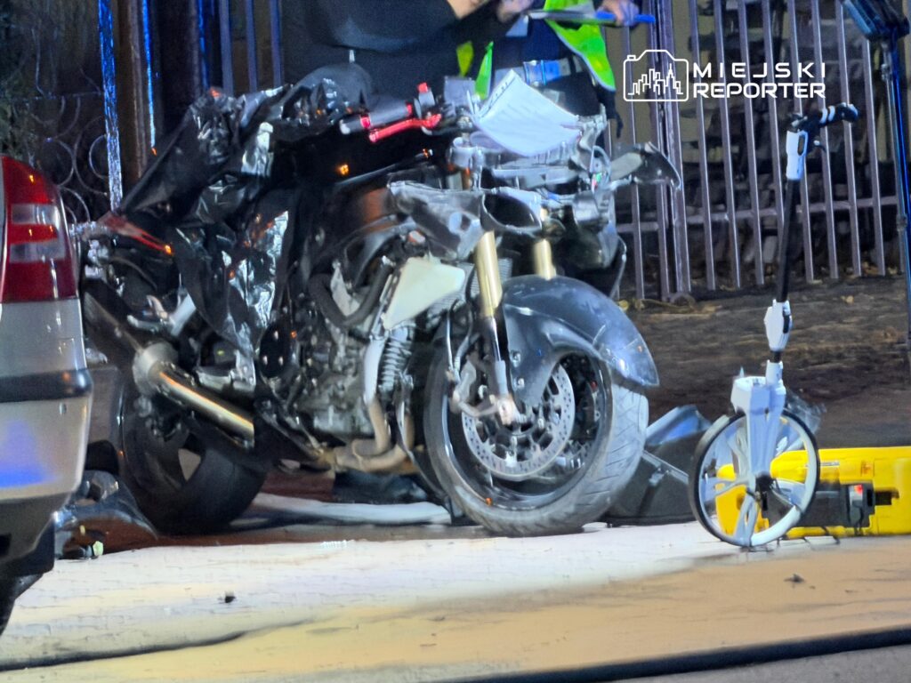 Uszkodzony motocykl stoi nocą przy barierkach, obok samochodu i żółtego sprzętu drogowego.