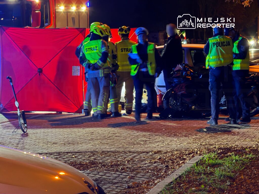 Strażacy i policjanci pracują nocą przy rozbitym samochodzie, obok którego ustawiono czerwoną osłonę.