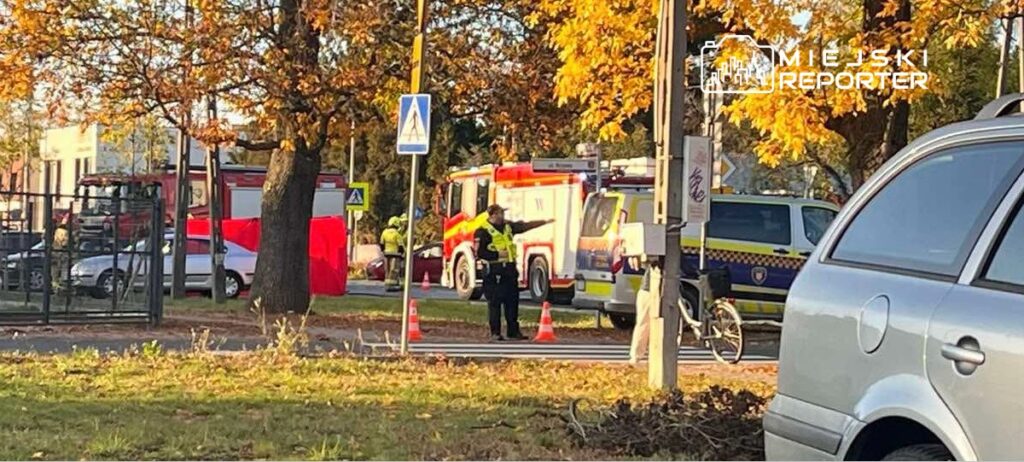 Strażacy i policjanci zabezpieczają miejsce zdarzenia przy ulicy, obok wozu strażackiego i radiowozu, wśród jesiennych drzew.