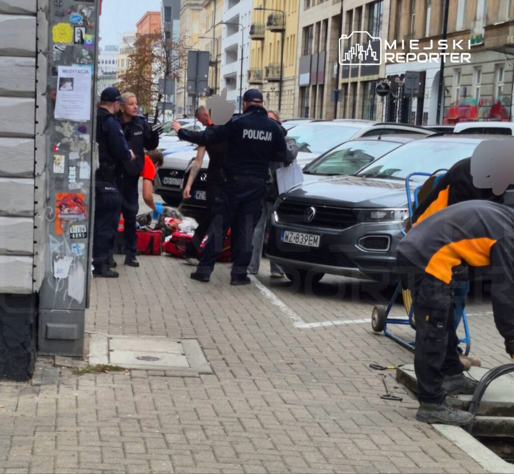 Policjanci interweniują przy zaparkowanych samochodach na miejskim chodniku, obok pracownika w odblaskowej odzieży.