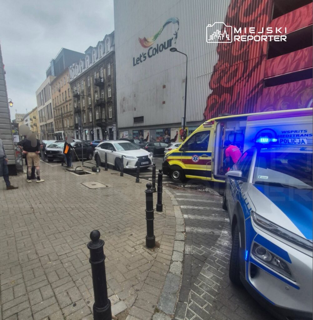 Radiowóz i żółty ambulans stoją przy ulicy, obok zaparkowanych samochodów i kamienic.