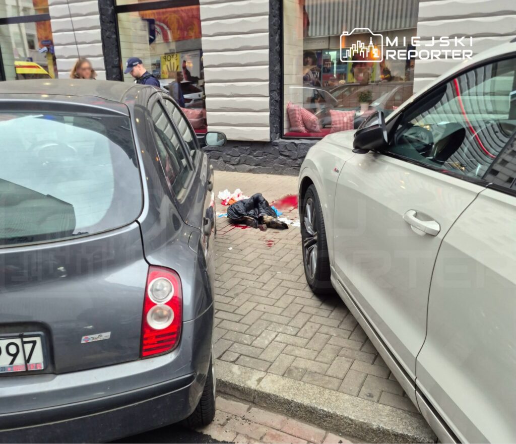 Osoba leży na chodniku między dwoma samochodami; wokół niej widać krew i porozrzucane przedmioty.