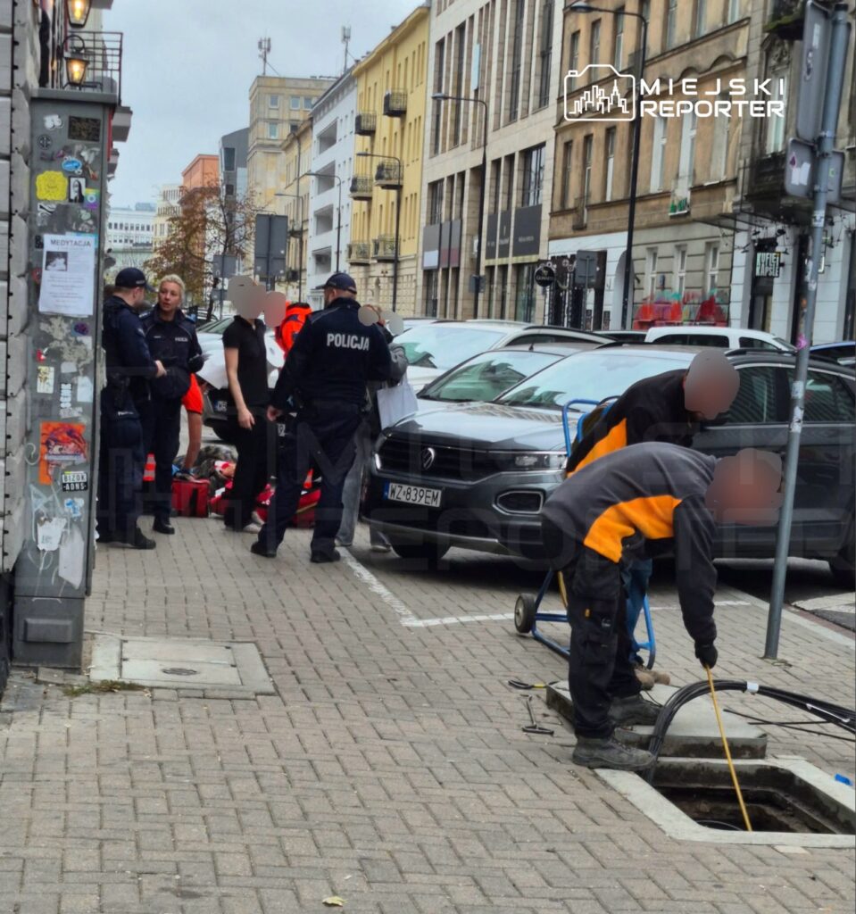 Policjanci i pracownicy w odblaskowych ubraniach przy uszkodzonym chodniku i zaparkowanych samochodach na miejskiej ulicy.