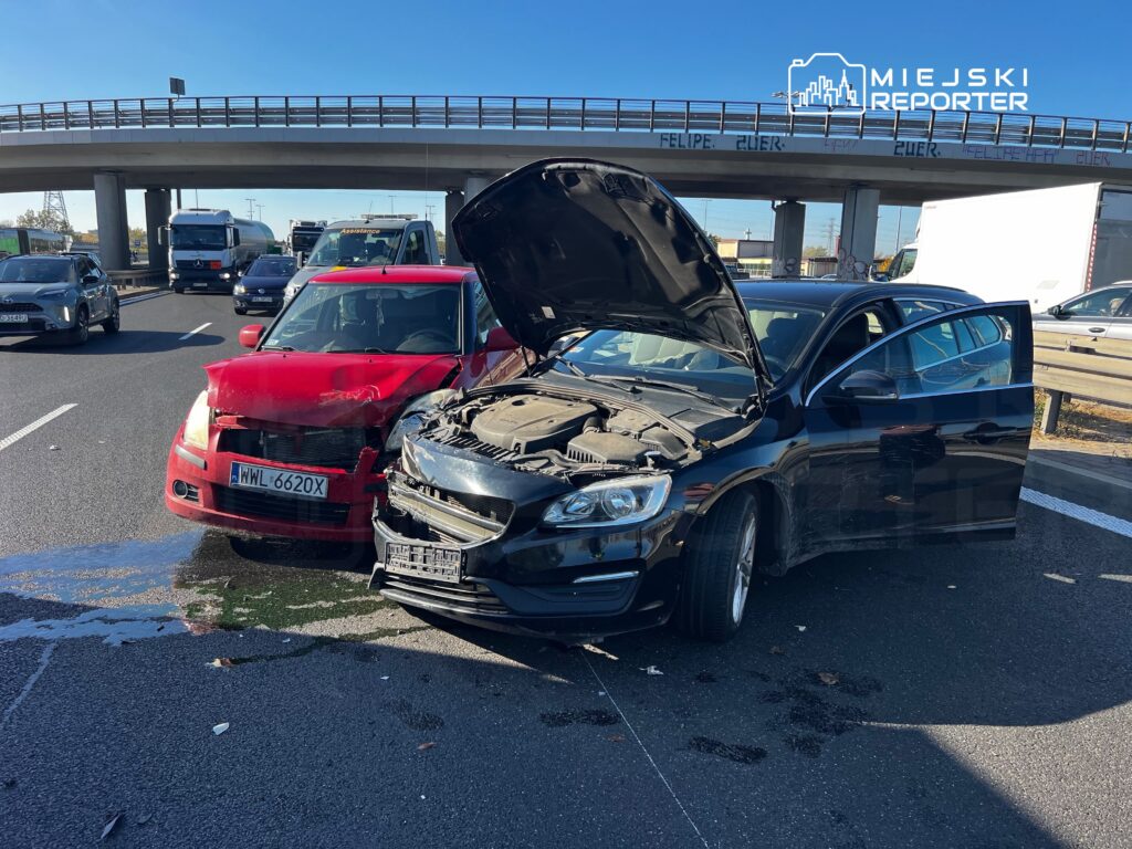 Uszkodzony czarny samochód z otwartą maską stoi przy czerwonym aucie na ruchliwej drodze pod wiaduktem.
