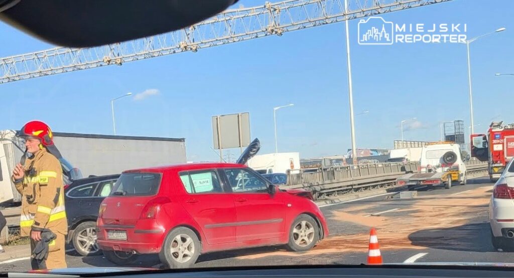 Strażak w hełmie stoi obok czerwonego samochodu na drodze, przy którym ustawiono pachołek ostrzegawczy.