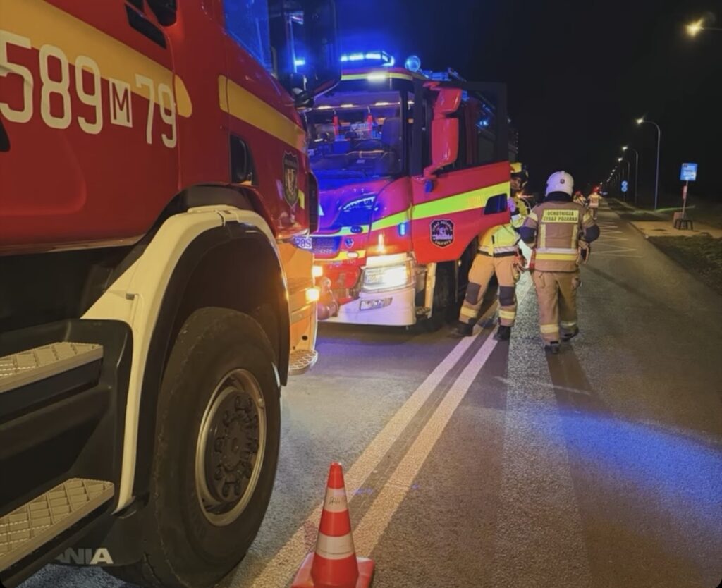 Strażacy w odblaskowych ubraniach pracują przy czerwonych wozach strażackich na nocnej drodze zabezpieczonej pachołkiem.