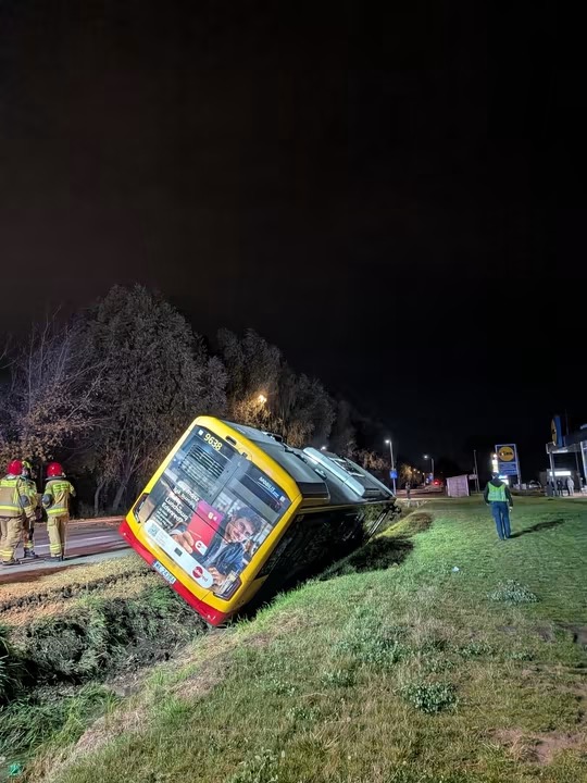 Żółty autobus leży przewrócony w przydrożnym rowie, a obok stoją służby ratunkowe nocą.