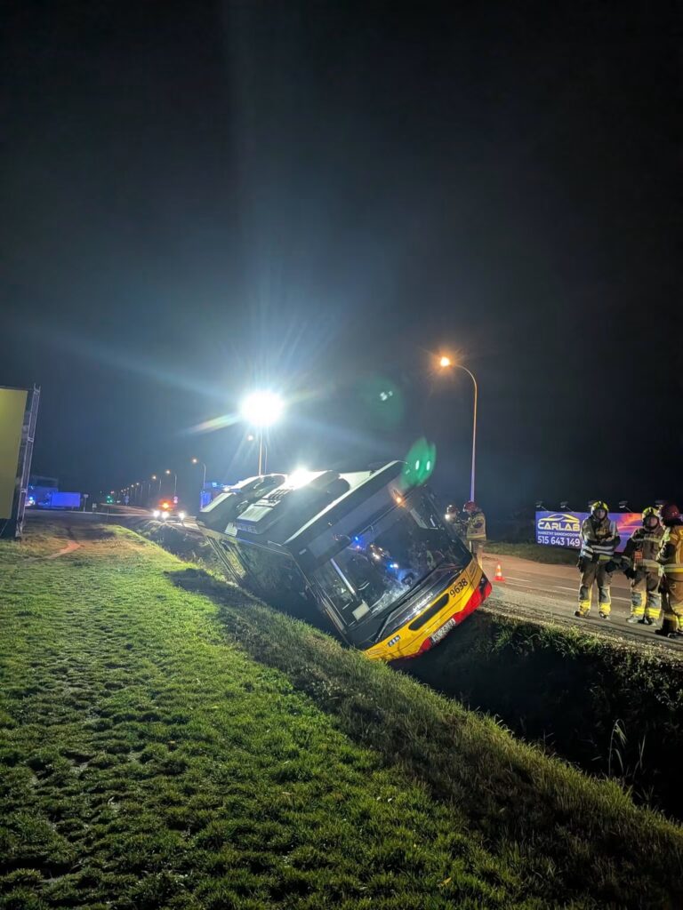 Autobus leżący na boku w przydrożnym rowie nocą, obok stoją strażacy i służby ratunkowe.