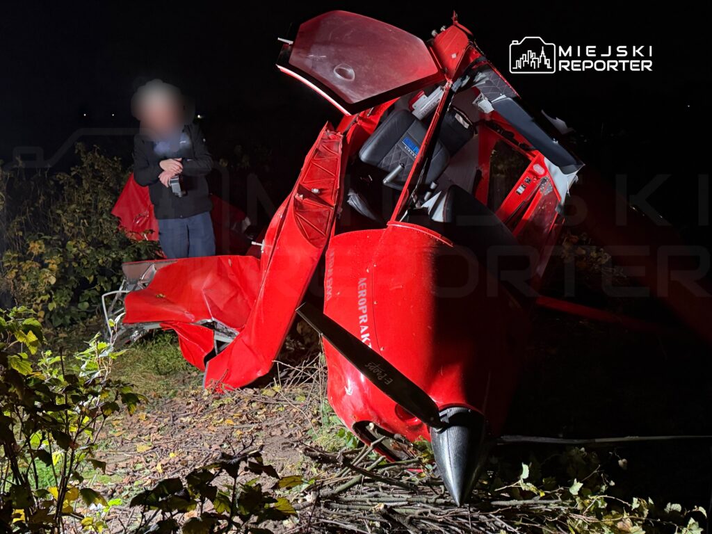 Rozbity czerwony śmigłowiec leży w zaroślach nocą, obok stoi osoba w ciemnym ubraniu.