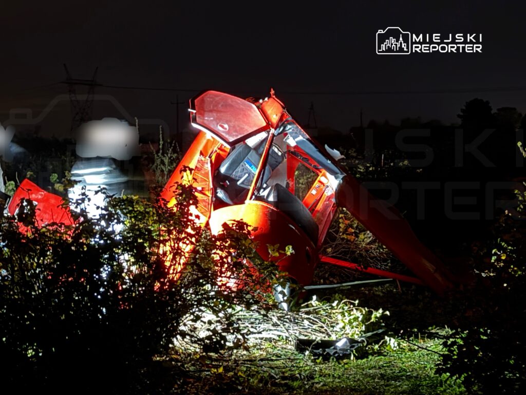 Rozbity czerwony pojazd leży w zaroślach nocą, oświetlony reflektorami służb ratunkowych.