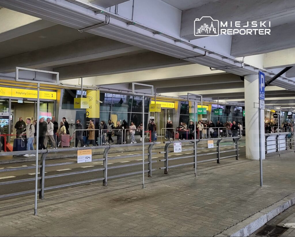 Pasażerowie stoją w kolejce przed przeszklonymi wejściami na lotnisko, pod żółtymi tablicami „Przyloty | Arrivals”.