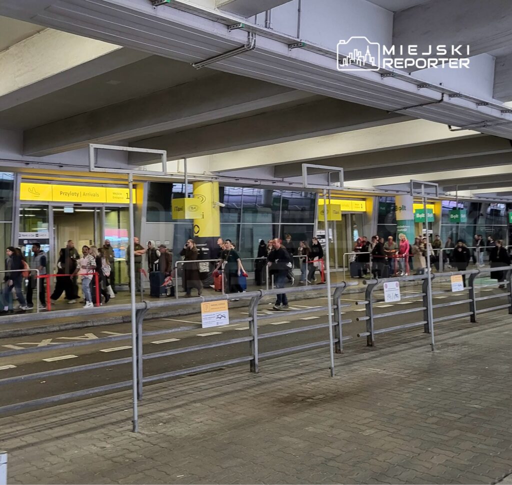 Pasażerowie stoją przy stanowiskach odbioru podróżnych pod żółtym szyldem „Przyloty / Arrivals” na lotnisku.