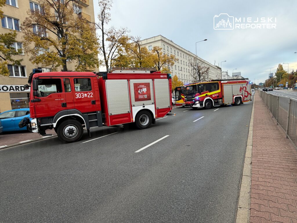 Dwa czerwone wozy strażackie stoją na miejskiej ulicy przy chodniku i budynkach.