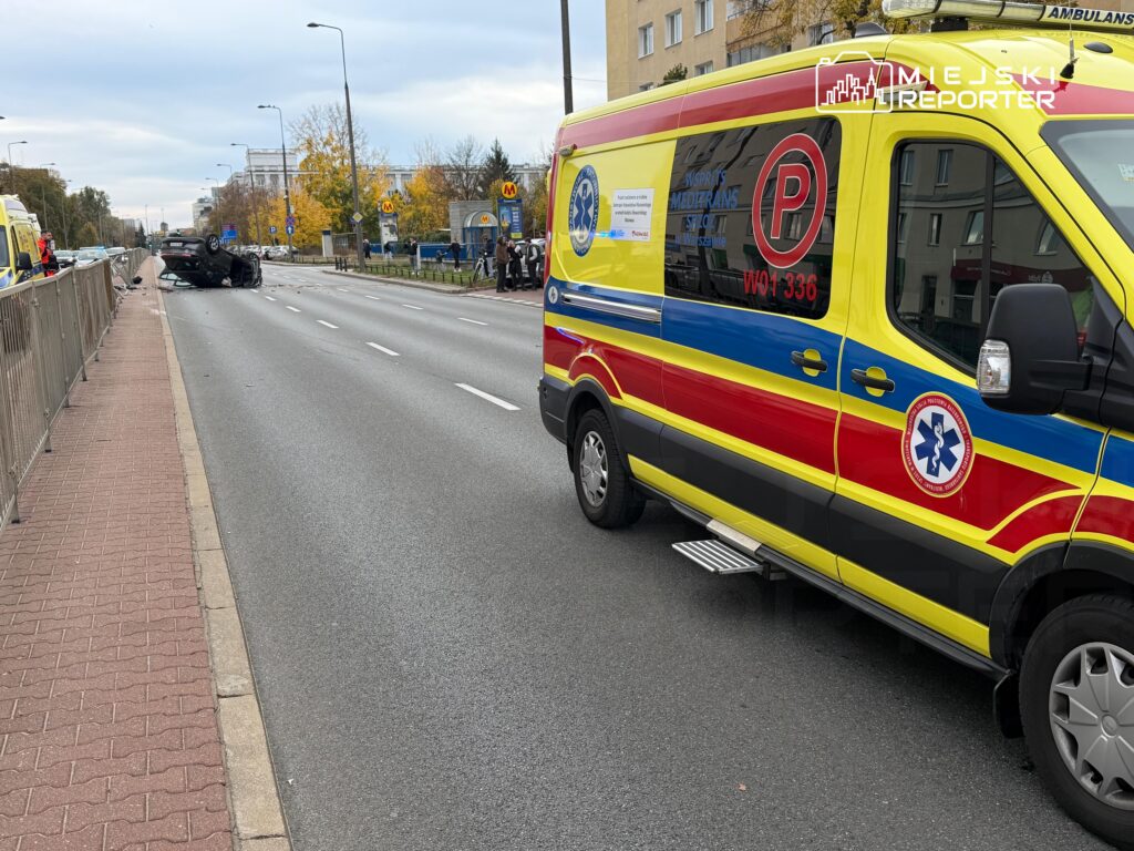 Żółto-czerwony ambulans stoi na jezdni, a dalej widać rozbity samochód i służby ratunkowe.