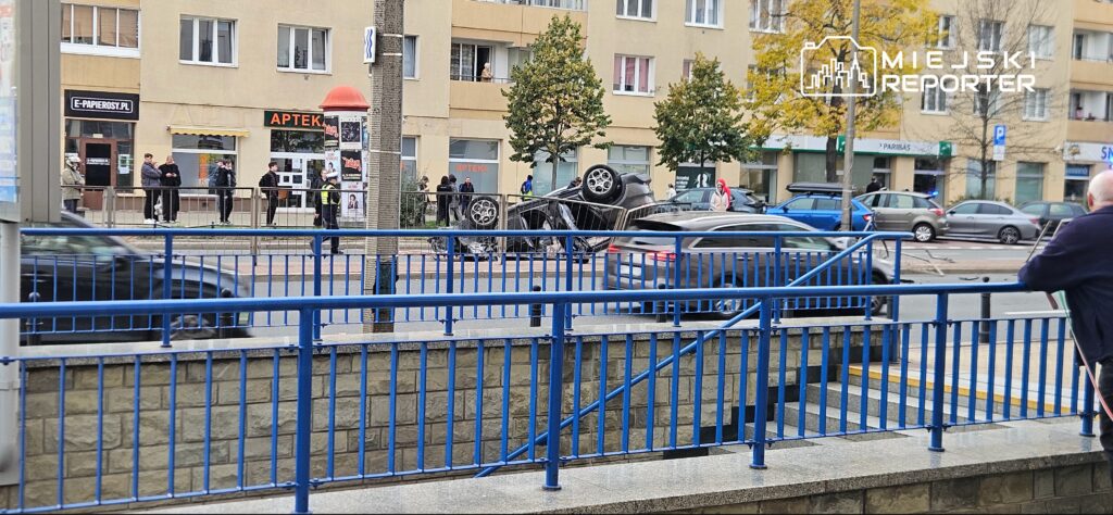Samochód leży na dachu na jezdni, za niebieskimi barierkami, przed budynkiem z lokalami usługowymi.