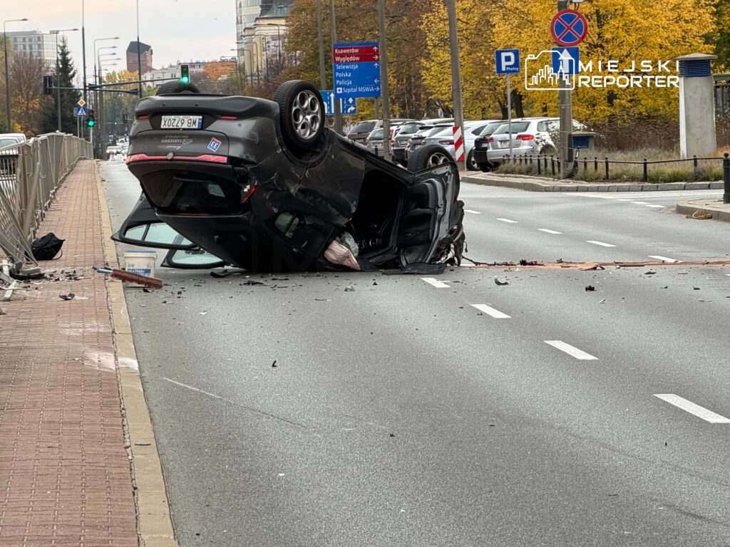 Czarny samochód leży na dachu na środku jezdni, wokół niego widać rozrzucone odłamki.