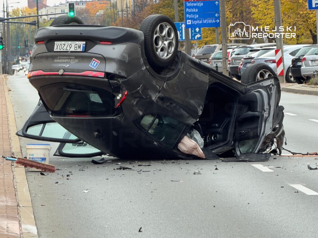 Ciemny samochód leży do góry kołami na jezdni po wypadku; wokół niego widać rozrzucone odłamki i stojące auta.