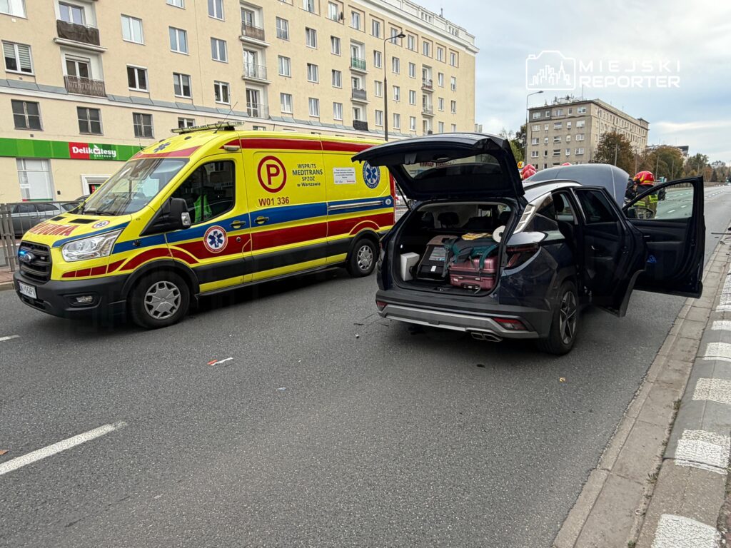 Żółty ambulans stoi obok czarnego SUV-a z otwartym bagażnikiem i drzwiami na ulicy przy chodniku.