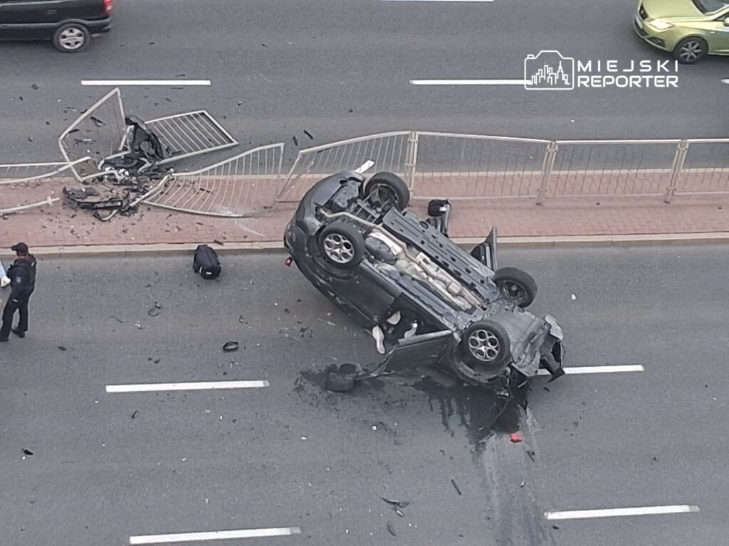 Samochód leży na dachu na jezdni, obok uszkodzonego ogrodzenia i rozrzuconych szczątków.