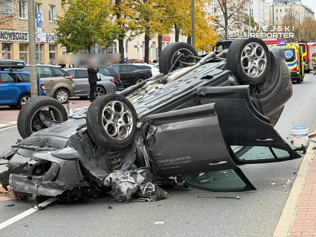 Rozbity samochód leży na dachu na miejskiej jezdni, a za nim stoi karetka pogotowia.