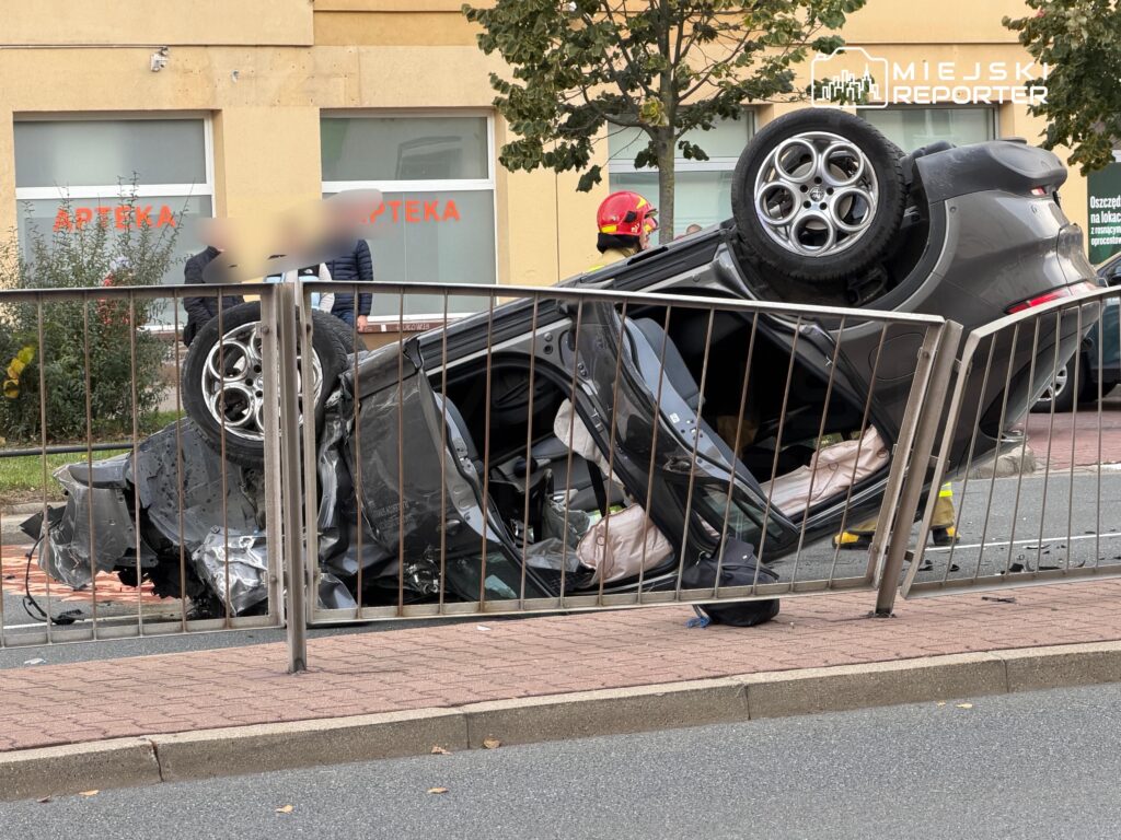Rozbity samochód leży na dachu za metalową barierką, przed budynkiem z szyldem „APTEKA”; obok stoi strażak.