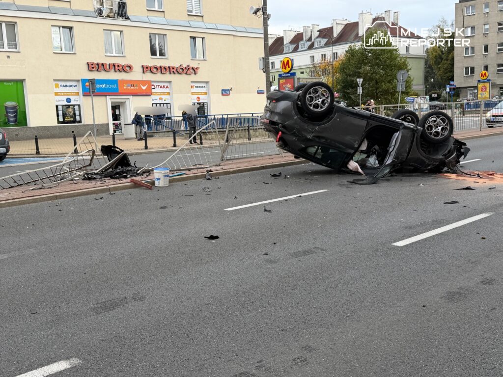Przewrócony na dach czarny samochód leży na jezdni przy uszkodzonych barierkach, a wokół niego rozsypane są odłamki.