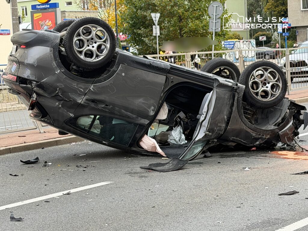 Rozbity szary samochód leży dachem na jezdni; wokół pojazdu widać odłamki i wyciek płynu.