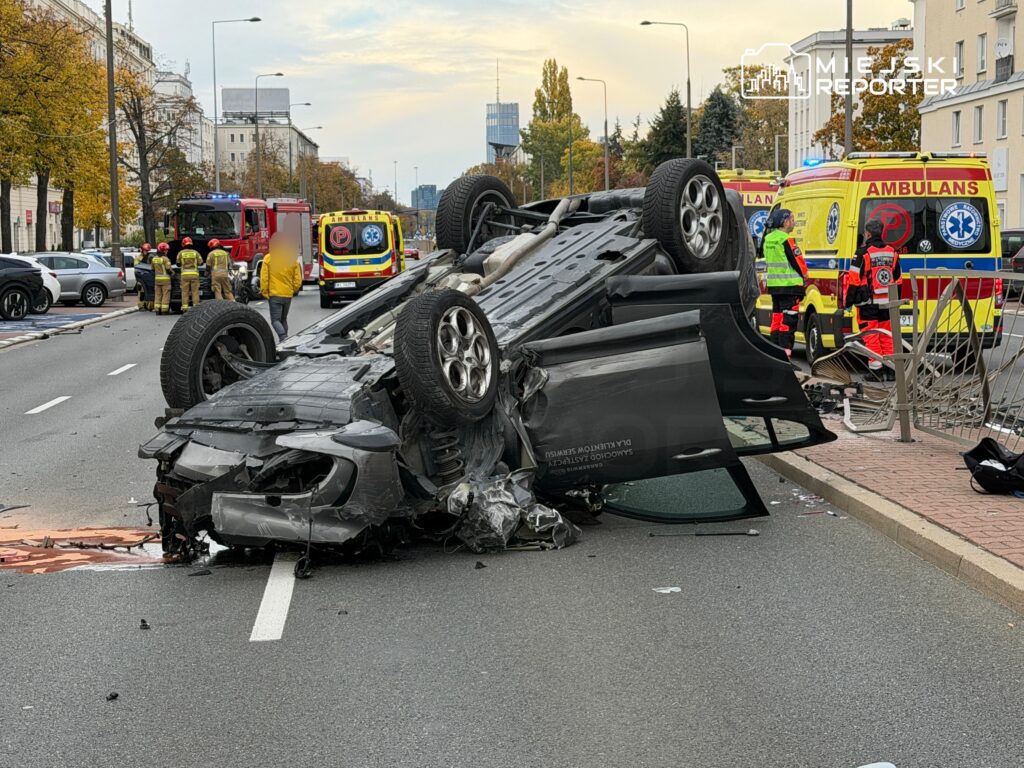 Rozbity samochód leży na dachu na środku ulicy, obok pracują strażacy i ratownicy medyczni.