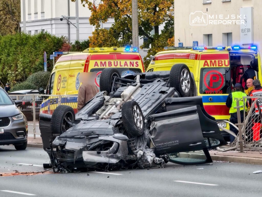 Przewrócony na dach, rozbity samochód leży na jezdni; obok stoją karetki i policjant.