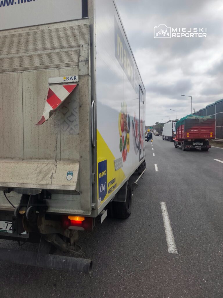 Ciężarówki stoją w kolejce na wielopasmowej drodze, za reklamowym samochodem dostawczym.