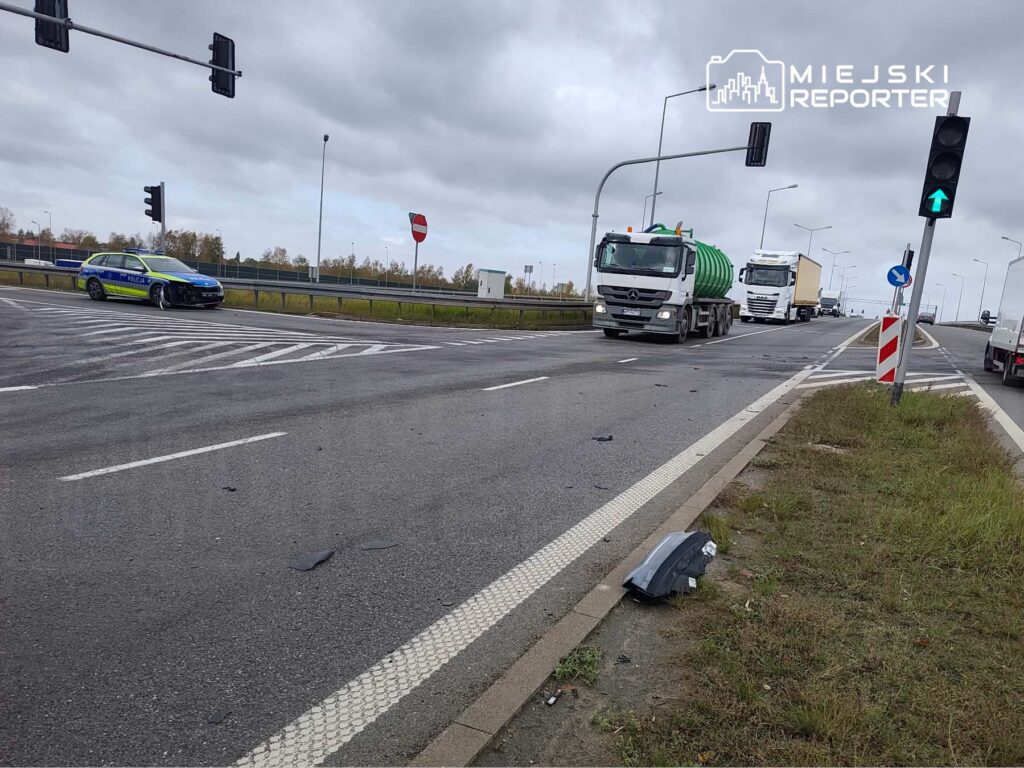 Ciężarówki przejeżdżają przez skrzyżowanie, obok stoi oznakowany radiowóz; na poboczu leżą fragmenty uszkodzonego pojazdu.