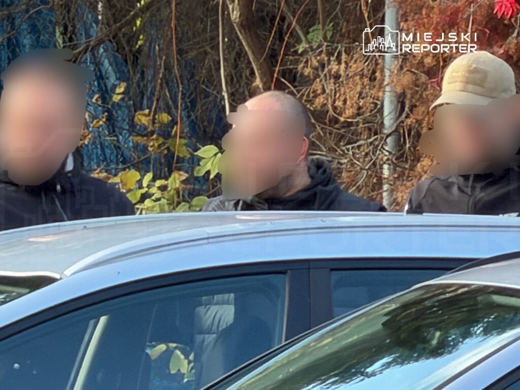 Trzech mężczyzn stoi za zaparkowanym samochodem na tle zarośli i drzew.