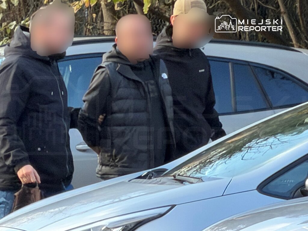 Trzech mężczyzn w ciemnych ubraniach stoi między zaparkowanymi samochodami.