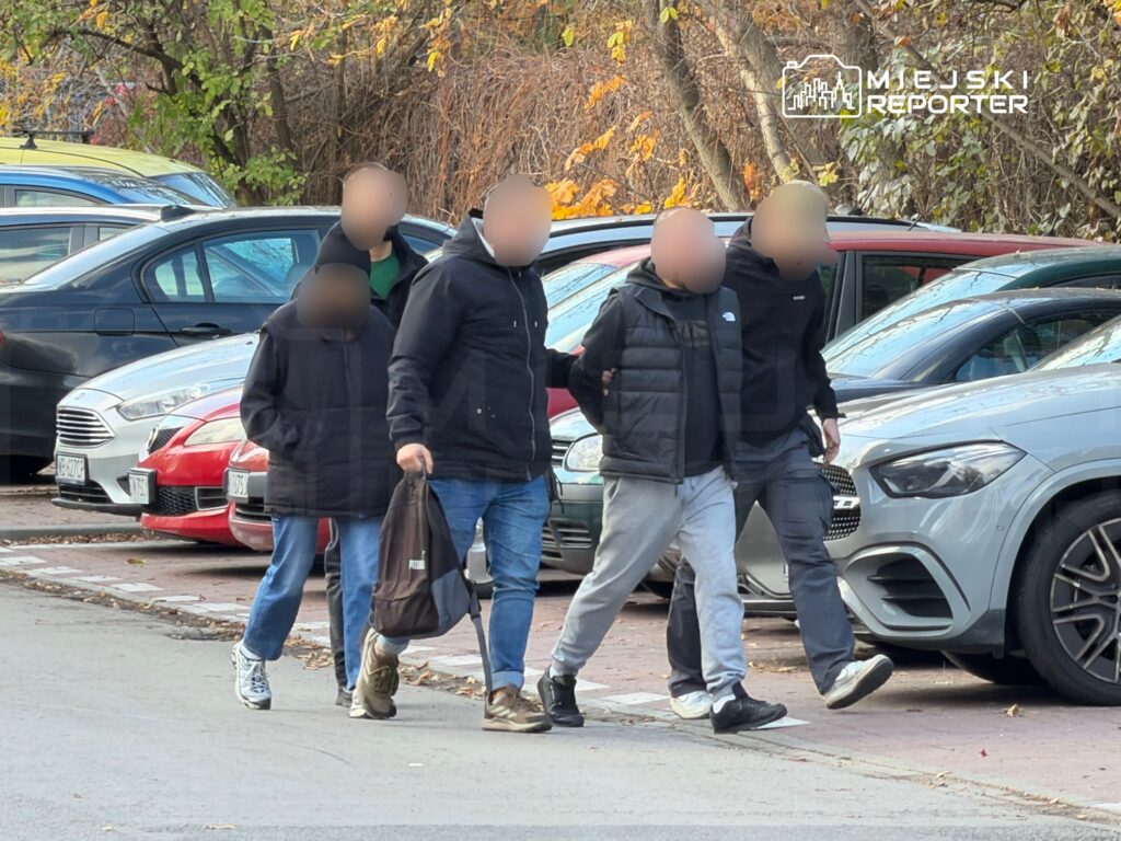 Pięciu mężczyzn idzie chodnikiem wśród zaparkowanych samochodów.