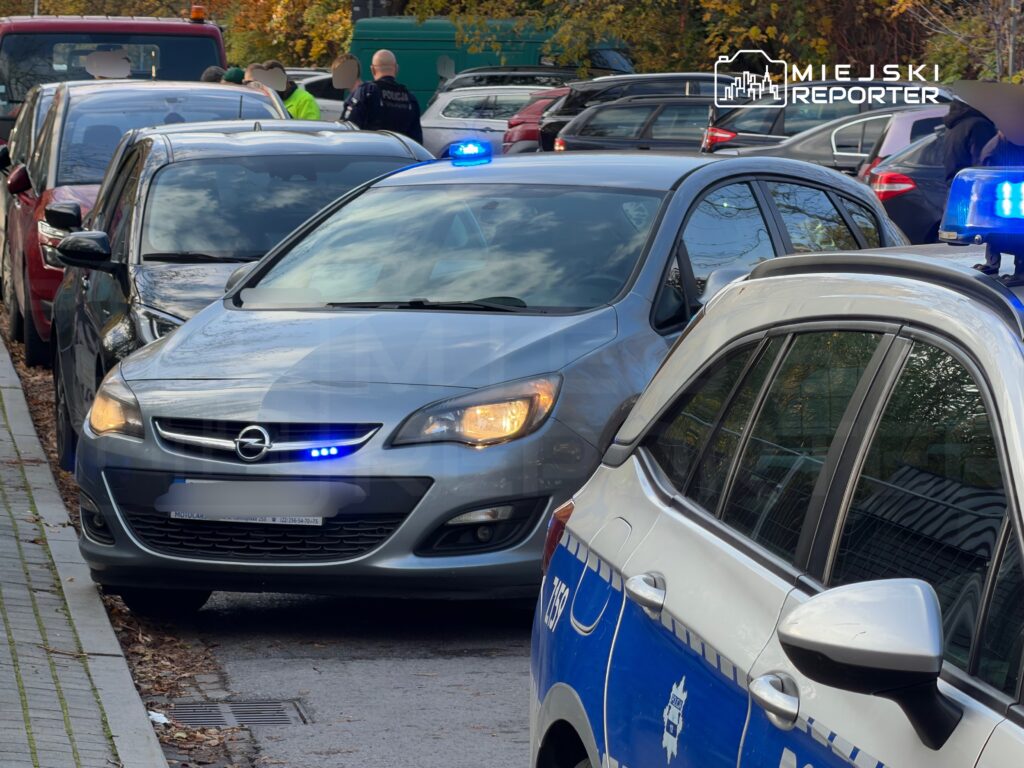 Szary Opel z włączonymi niebieskimi światłami stoi wśród samochodów obok oznakowanego radiowozu i funkcjonariuszy.