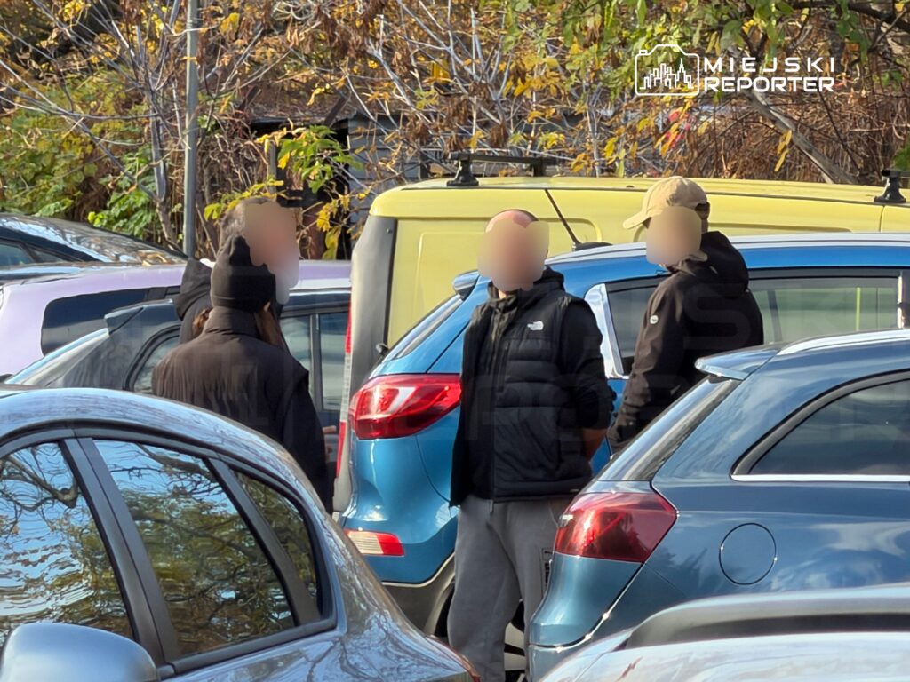 Trzy osoby rozmawiają między zaparkowanymi samochodami na parkingu, w tle widać drzewa z jesiennymi liśćmi.