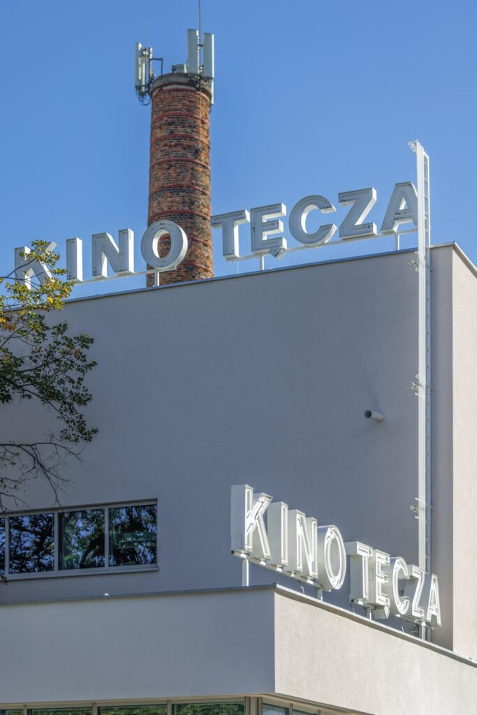 Napis „KINO TĘCZA” na elewacji jasnego budynku z ceglastym kominem na dachu, na tle błękitnego nieba.